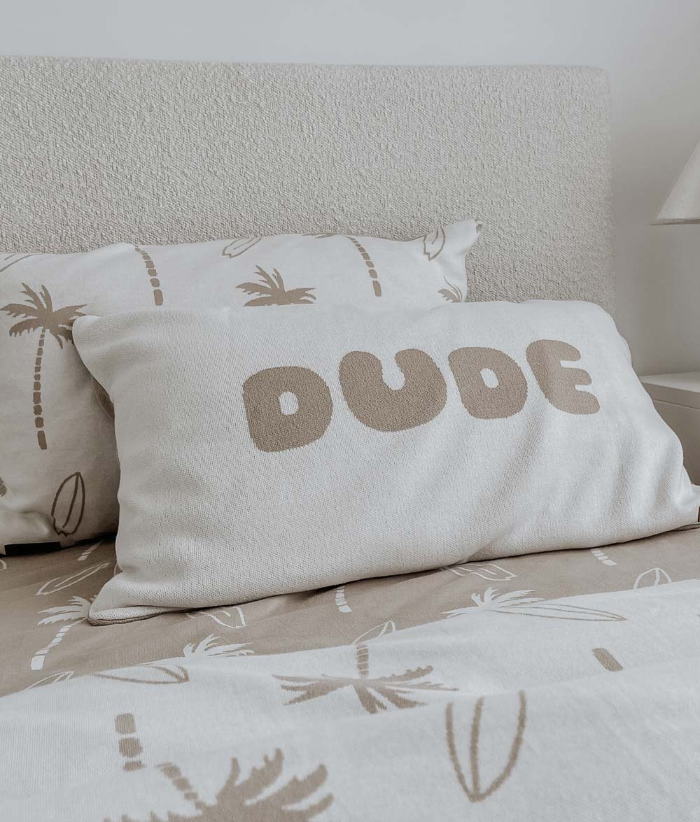 DUDE jersey cotton pillowcase layered in front of a Surfing Palm pillowcase on a sand fitted sheet