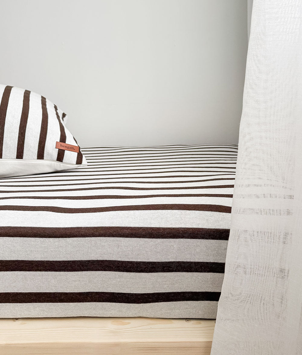 Side view of the Sundown Stripe fitted sheet on a timber bed beside a sheer ivory curtain