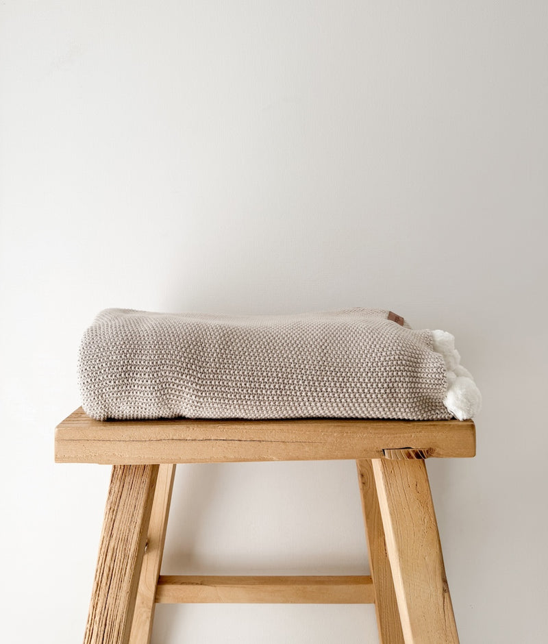Folded Greige Pom-Pom lounge throw with ivory pom poms on a rustic timber stool