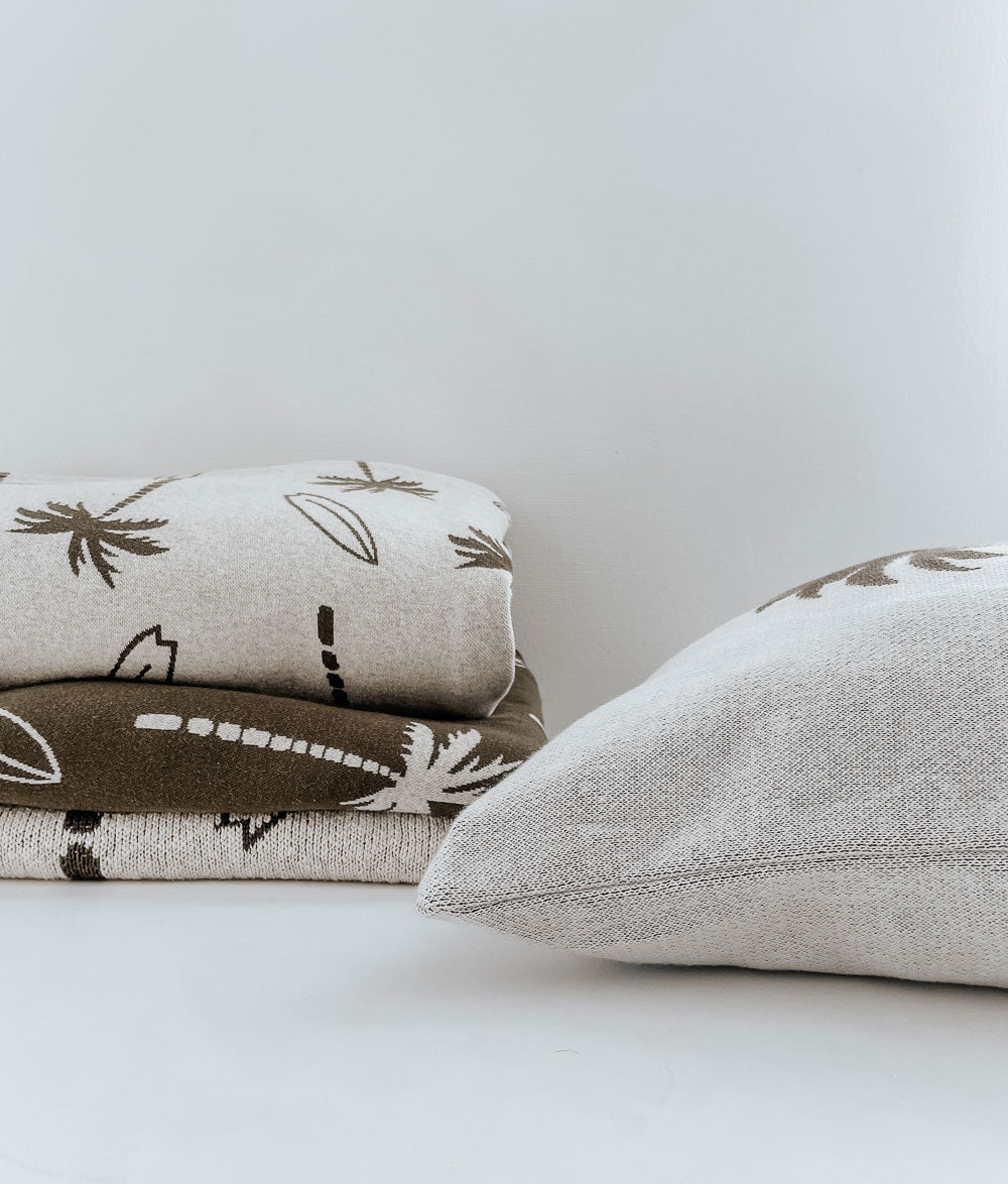 Folded Surfing Palm jersey fitted sheets in natural and olive stacked beside a plain ivory pillow
