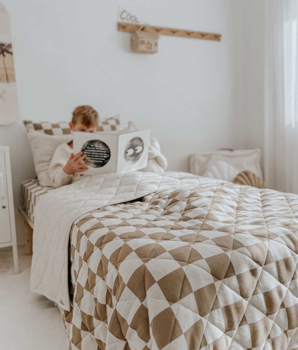 Child reading in bed with the khaki gingham quilt folded across the foot