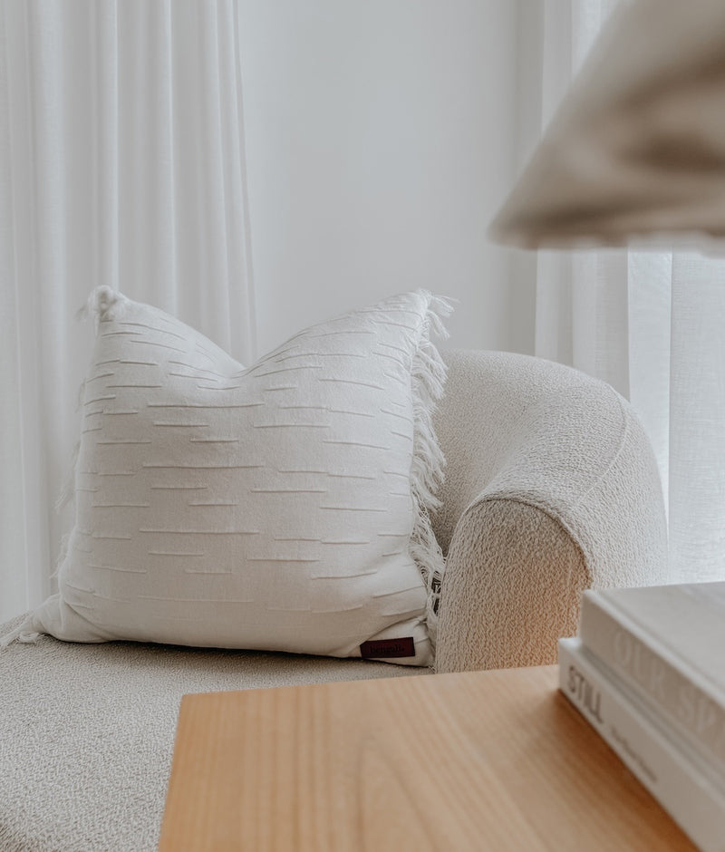 Ivory Flurry cushion cover with a fringed, textured finish on a boucle armchair beside a timber table