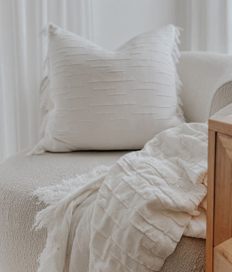 Ivory Flurry cushion cover and a fringed ivory throw on a boucle chair beside a timber side table