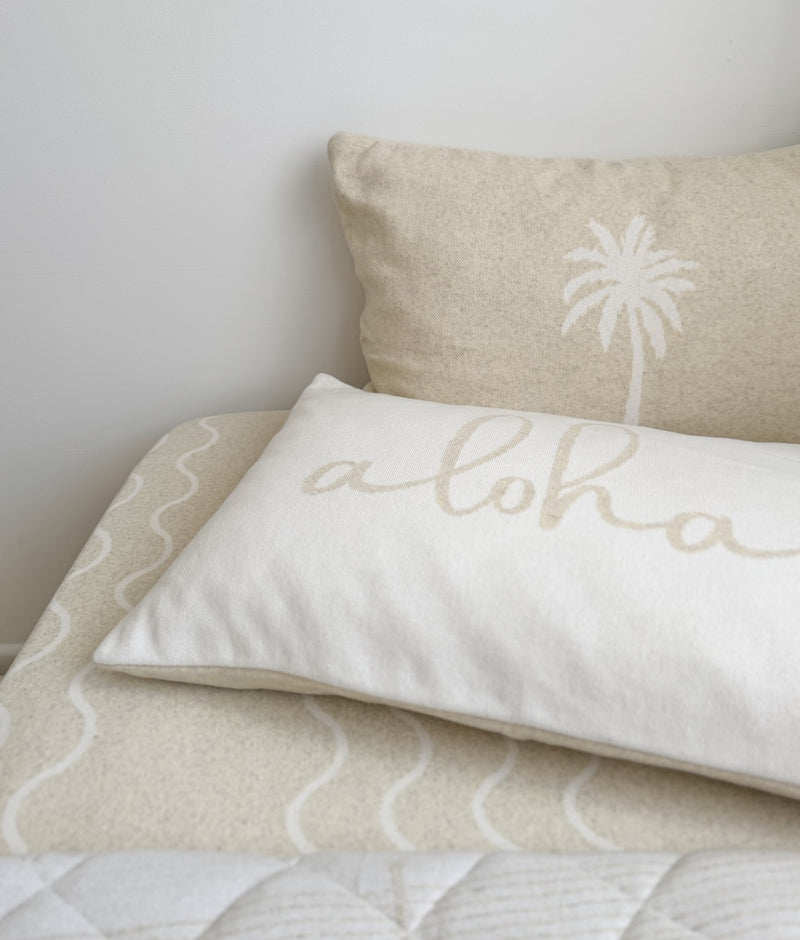 Aloha cotton pillowcase with natural script lettering in front of a palm pillowcase on a wave print sheet