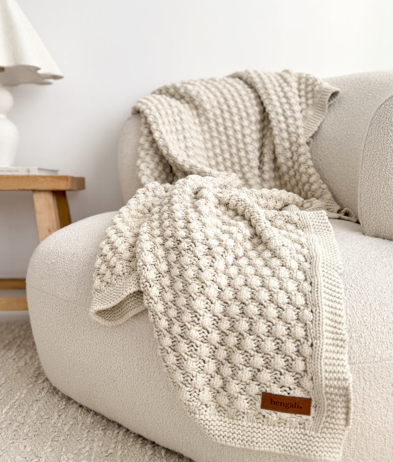 Bone popcorn-knit cotton lounge throw draped over a boucle armchair beside a pleated lamp and timber side table