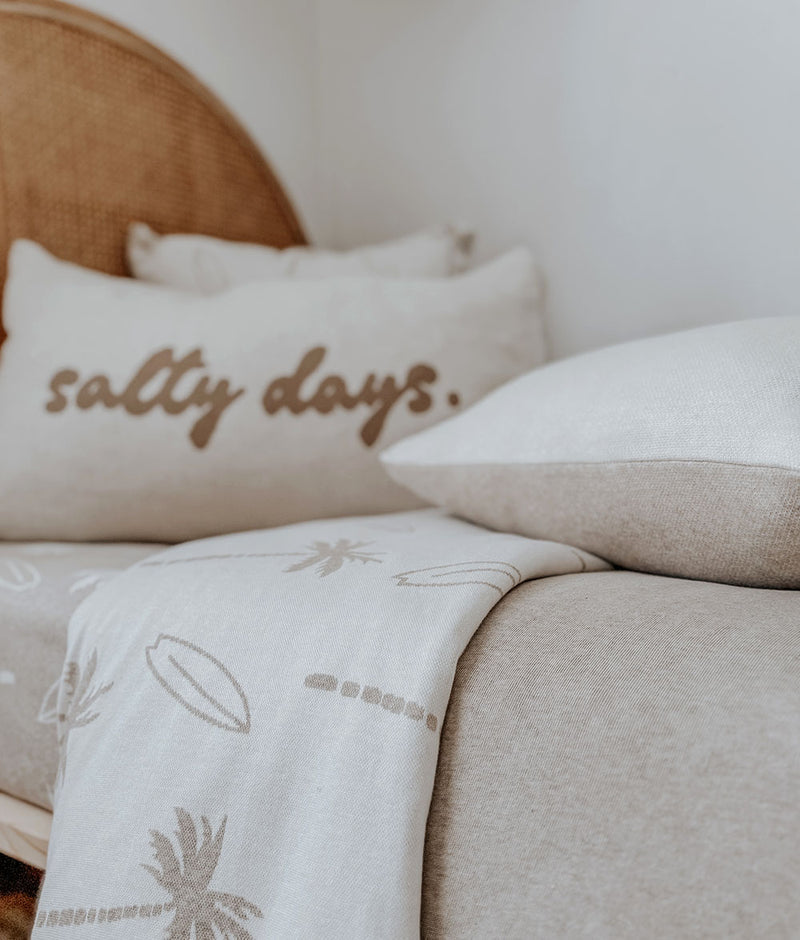 Salty Days pillowcase with a palm knit blanket on a sand bed beside a rattan bedhead