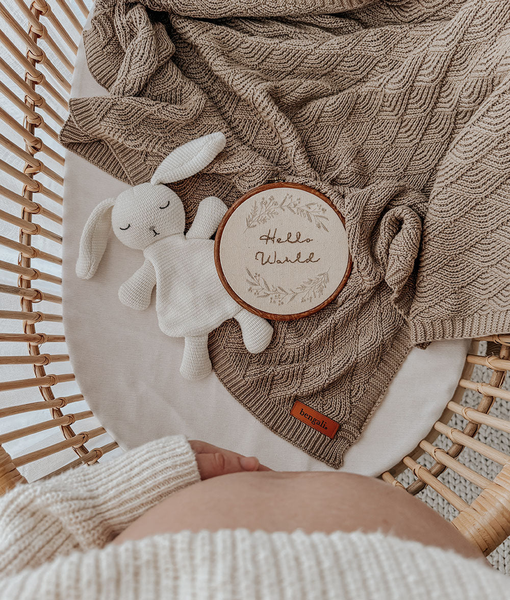 Overhead view of the stone blanket in a bassinet with a white bunny comforter and a Hello World disc