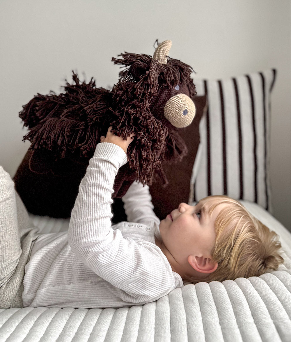 Child holding Fergus the Highland cow soft toy above them on a bed with a striped pillow