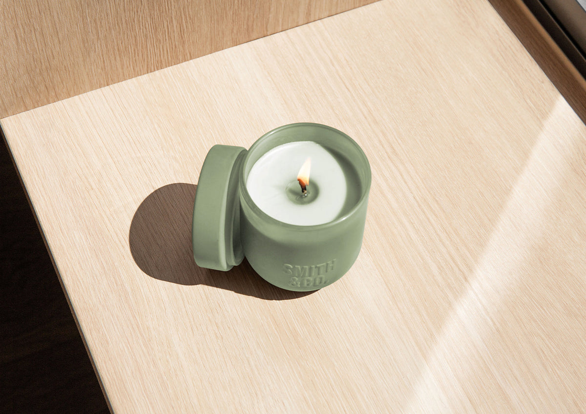 Lit Smith and Co candle with its lid beside it on a pale timber surface in sunlight