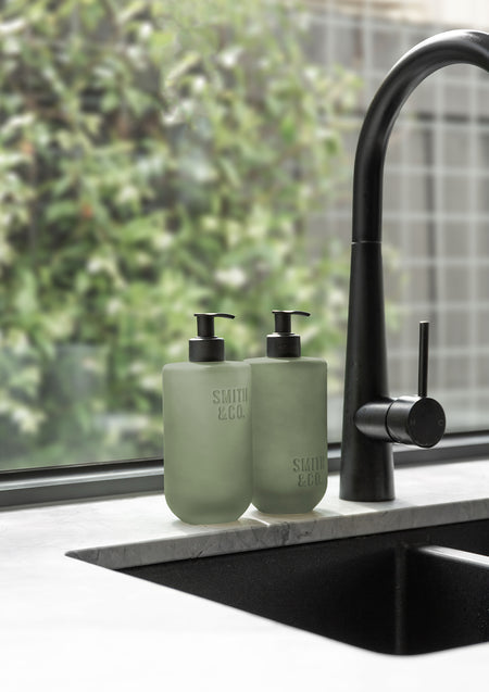 Sage green Smith & Co. bottles beside a black tap and sink, with a garden view behind