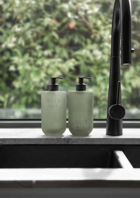Two sage green pump bottles by a black gooseneck tap in front of a leafy window