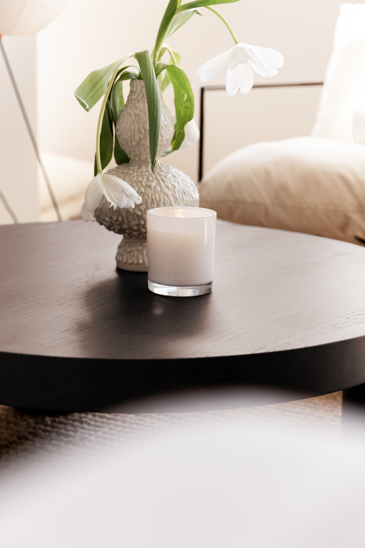 Lit Naturals candle beside a textured vase of white tulips on a dark round table