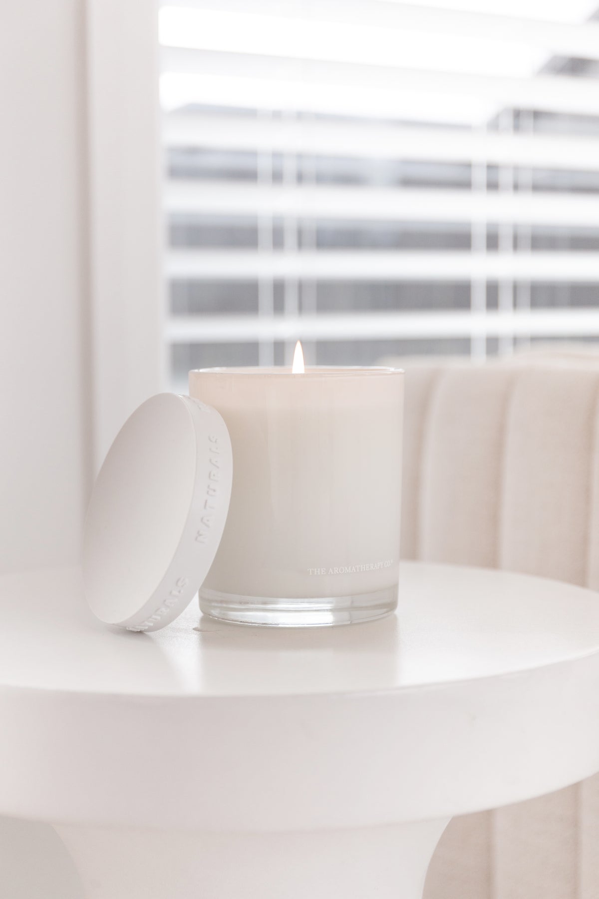 Lit Naturals candle with its lid propped on a white side table by a shuttered window