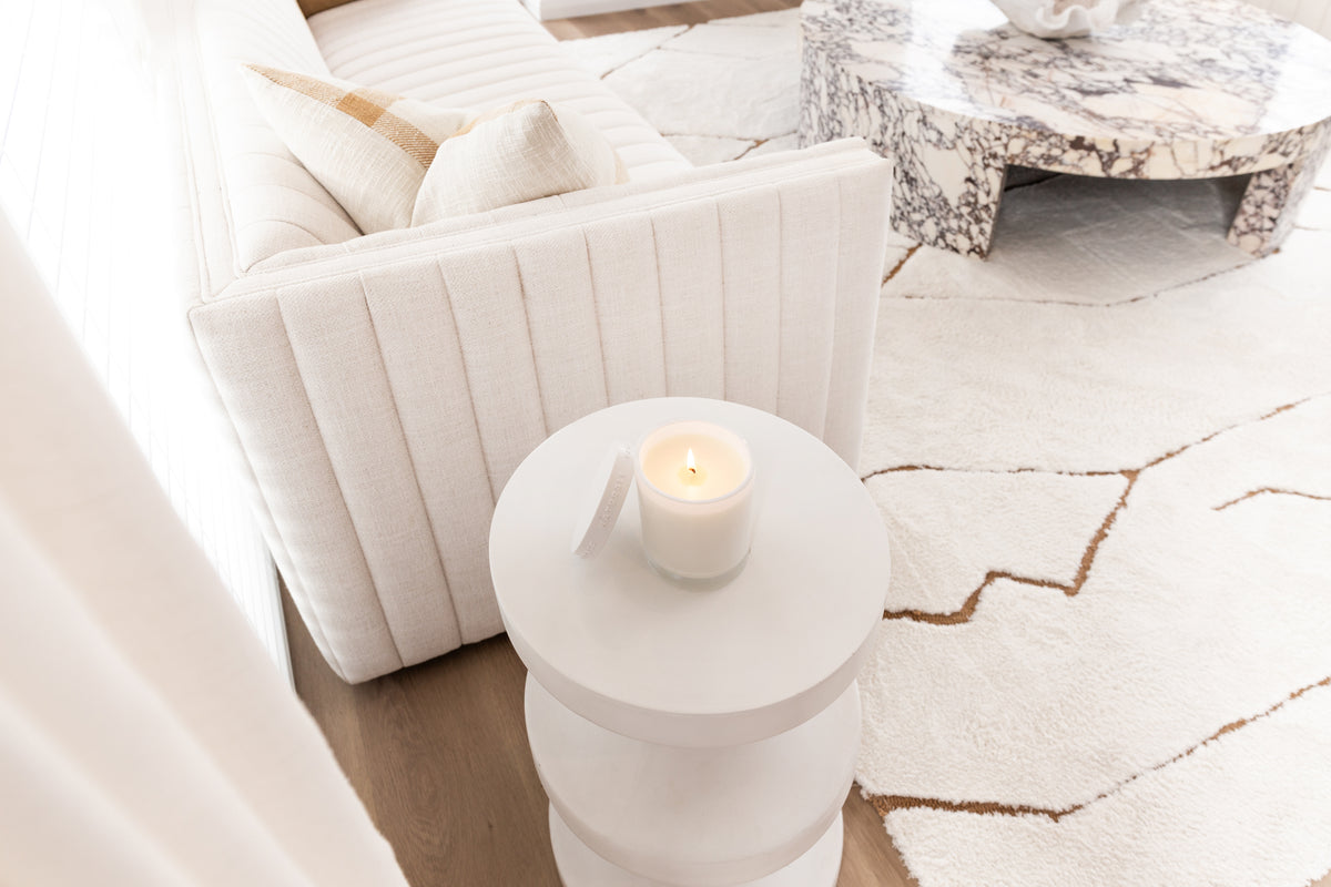 Lit Naturals candle with its lid on a white drum side table beside a fluted sofa