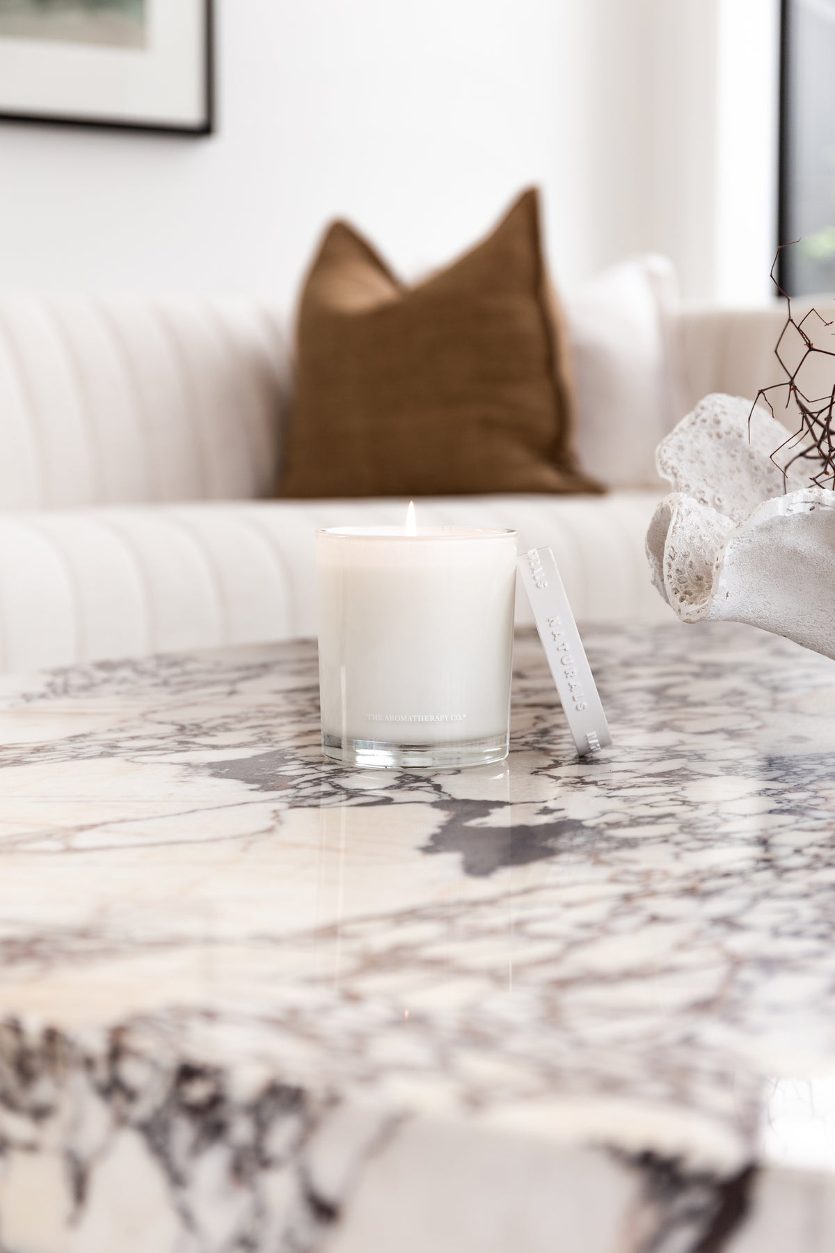 Lit Naturals candle and its lid on a veined marble table with a fluted sofa behind