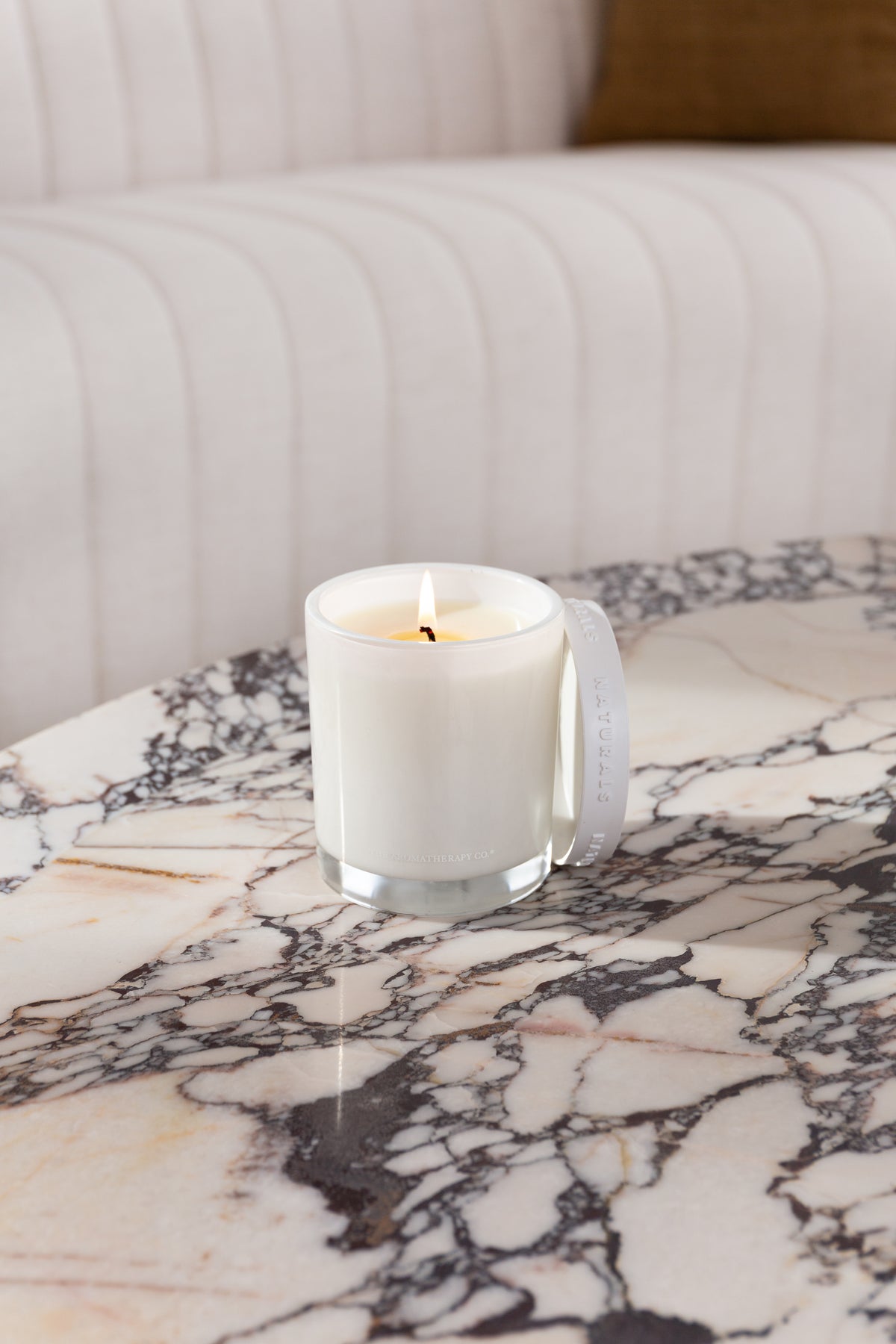 Lit Naturals candle and its lid on a veined marble tabletop in soft daylight