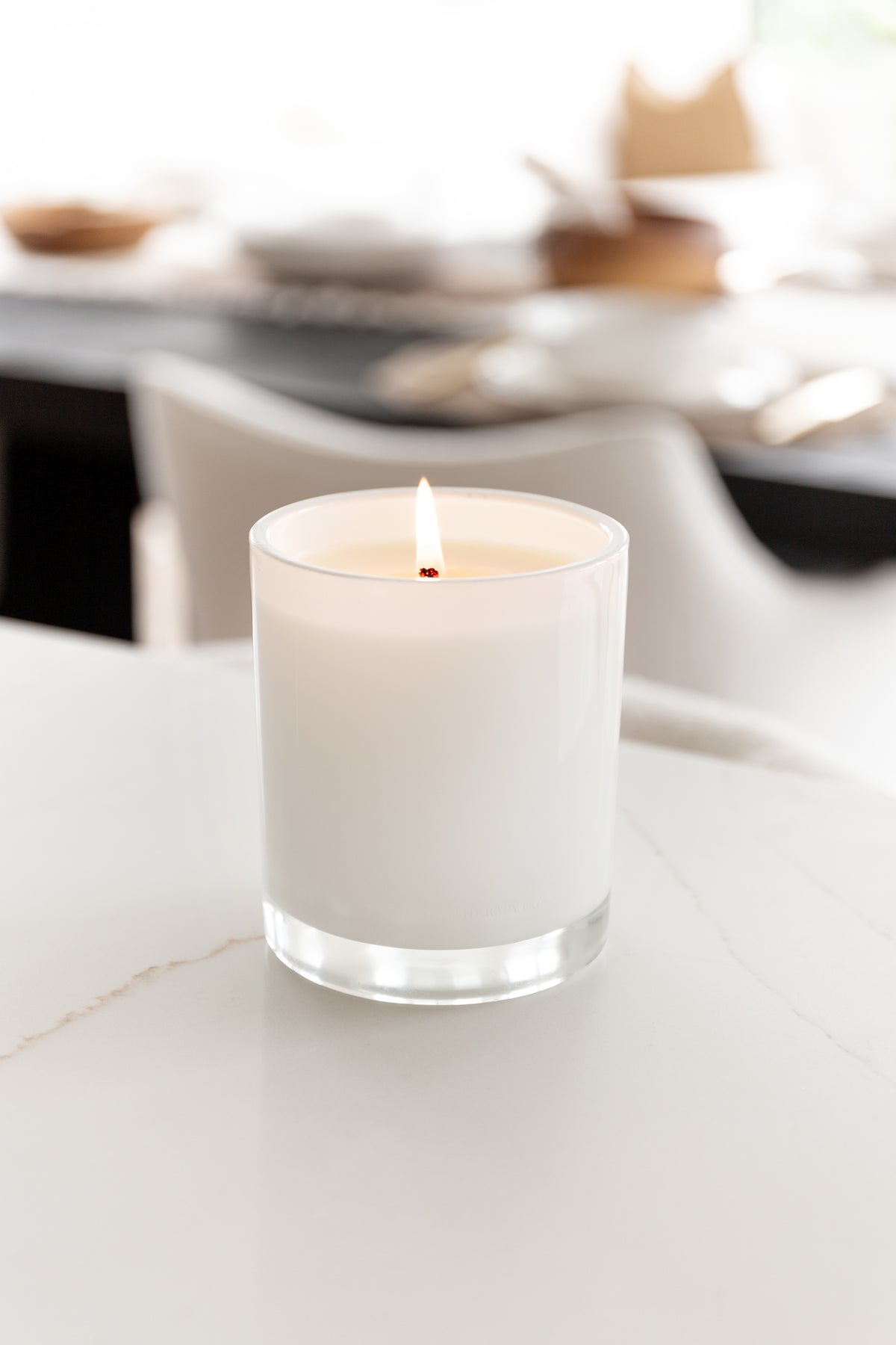 Lit Naturals candle on a white benchtop with the dining area softly out of focus