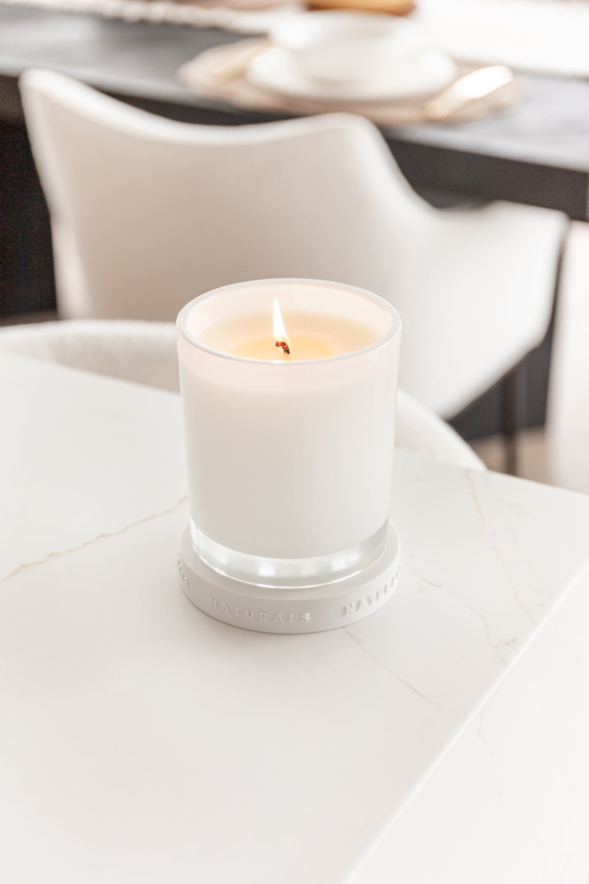 Lit Naturals candle on a white benchtop with dining chairs blurred behind