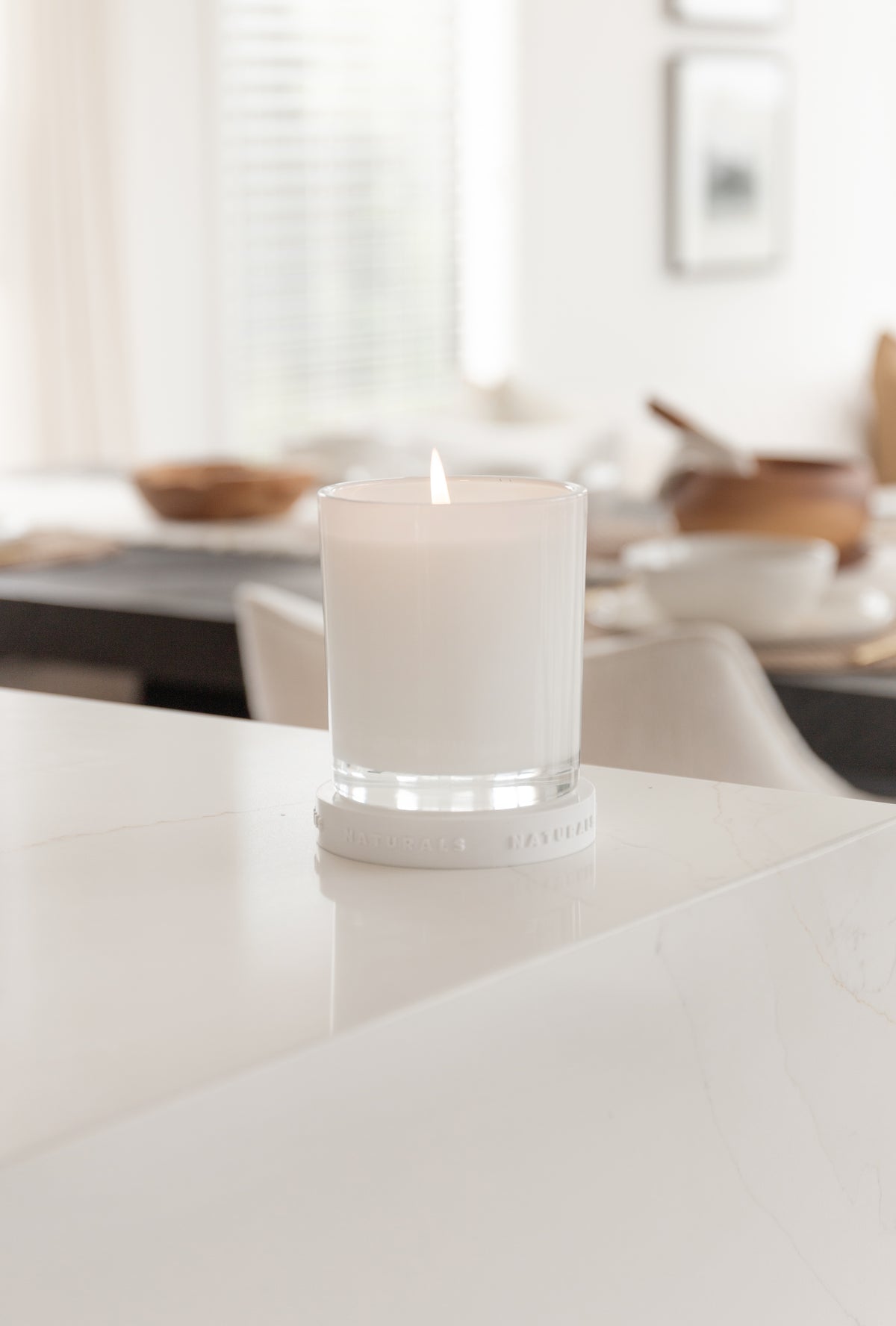 Lit Naturals candle on a white benchtop with a dining setting blurred behind