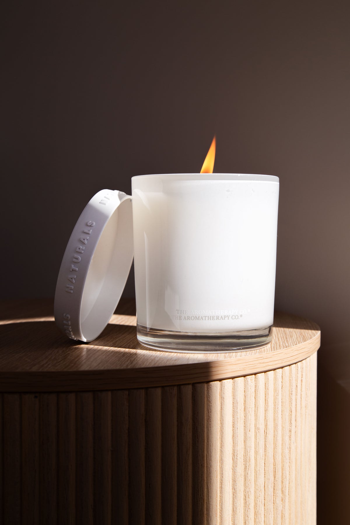 Lit Naturals candle with its lid propped against a fluted timber side table, dark wall behind