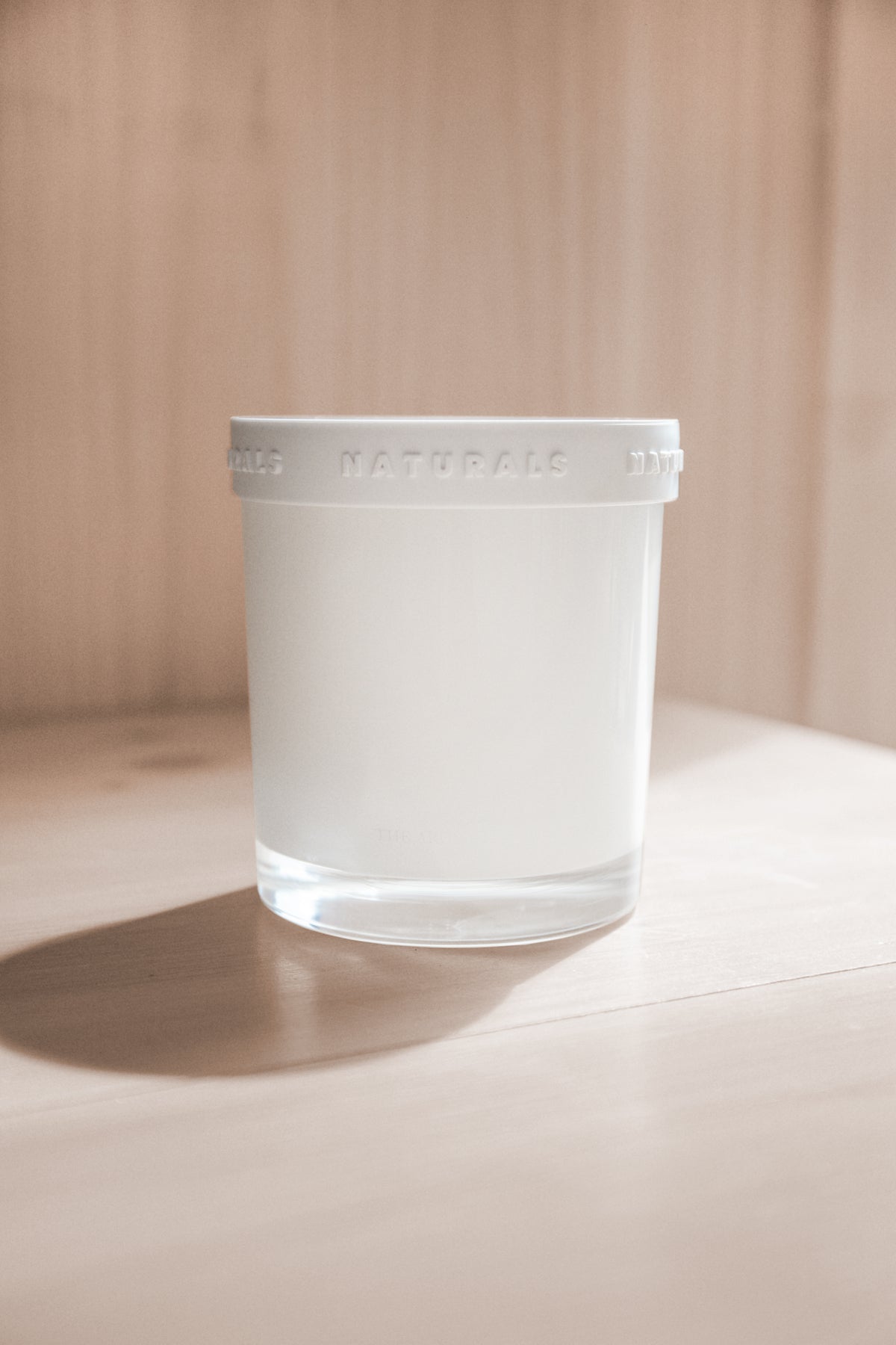 Naturals candle with its lid on, lit by hard sunlight against a pale timber wall