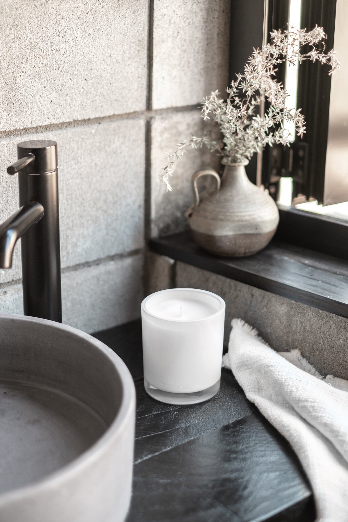 Naturals candle on a dark stone vanity beside a stone basin, black tap and dried stems