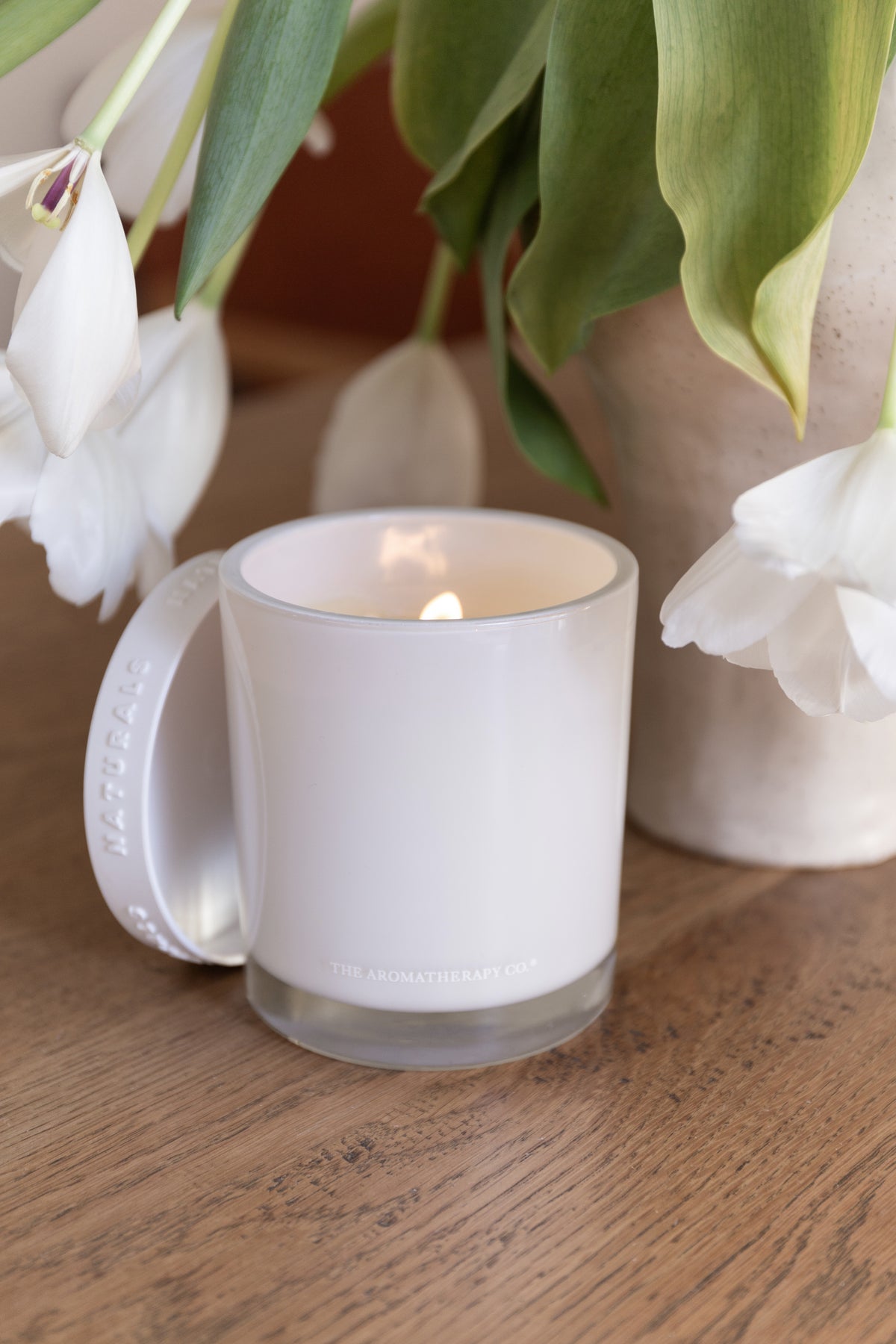 Close view of the lit Naturals candle beside white tulips on a timber table