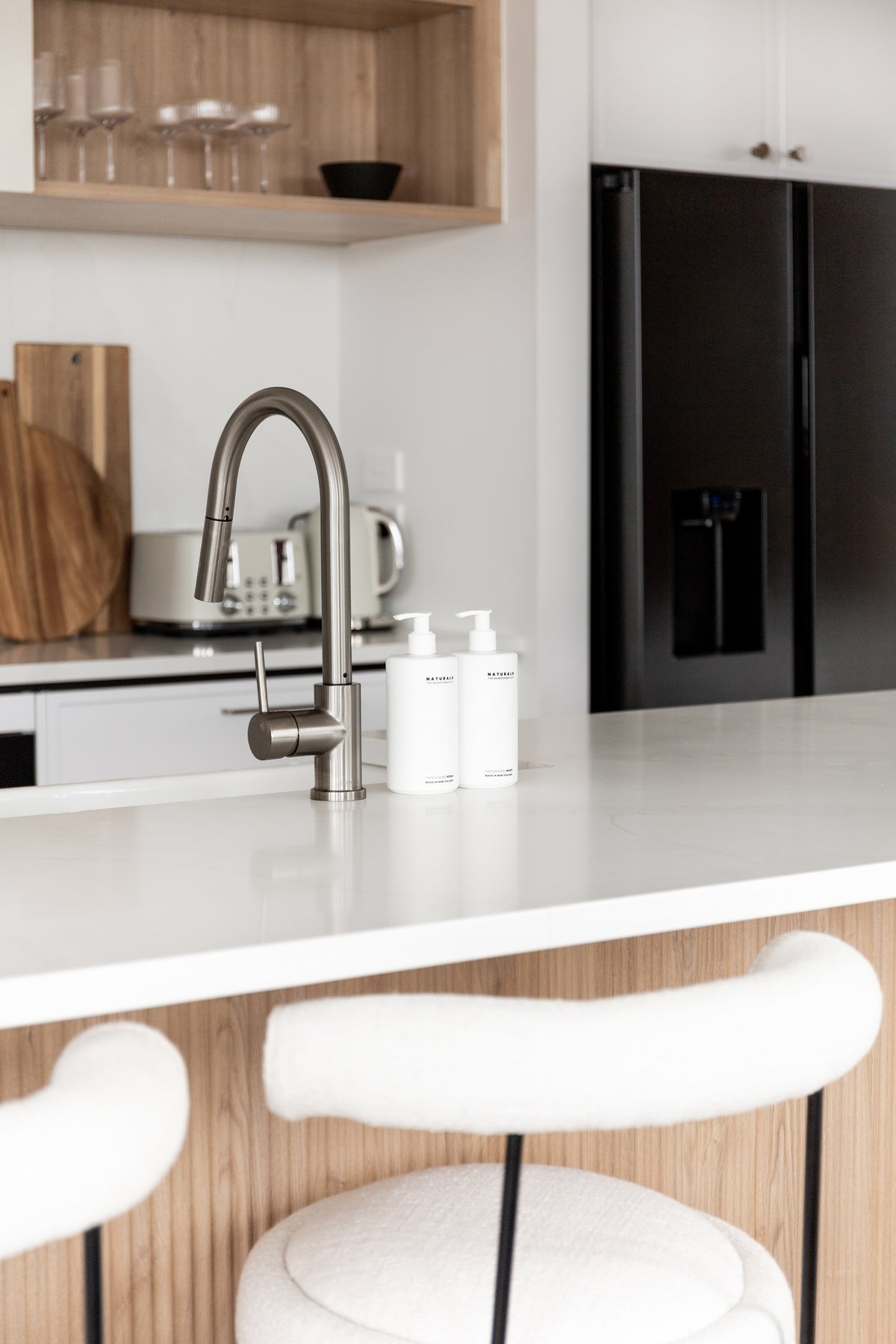 Naturals bottles beside a kitchen tap, the fluted timber island and white bar stools below