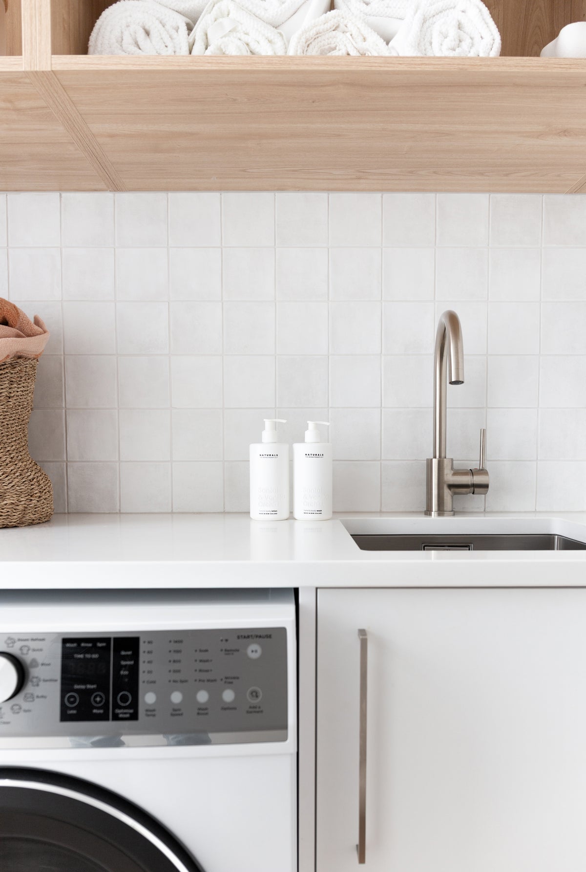 Square tiled laundry with rolled towels above, a front loader below and two Naturals pump bottles on the bench