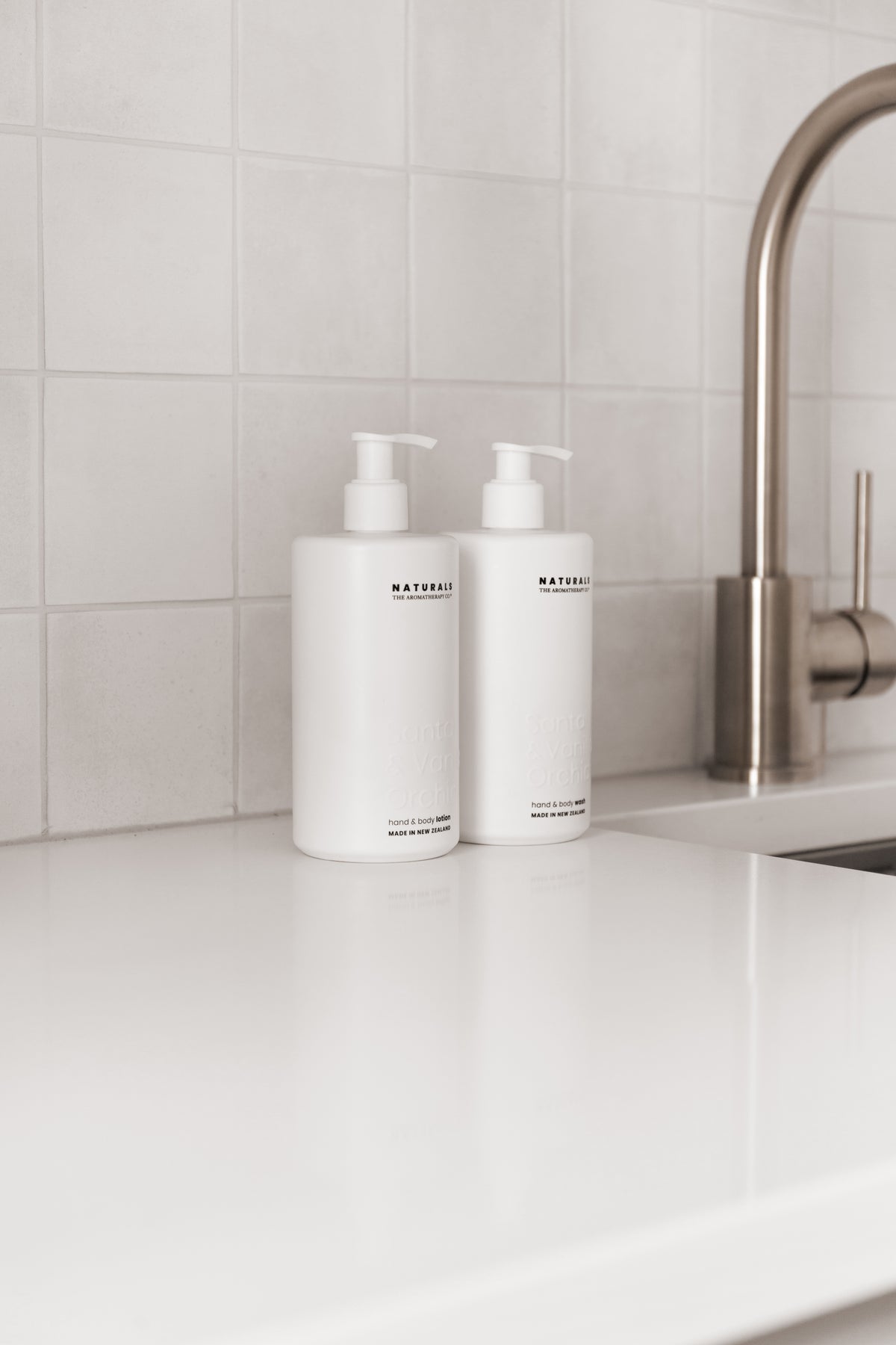 Two white Naturals pump bottles on a bench beside brushed tapware, square tiled splashback behind