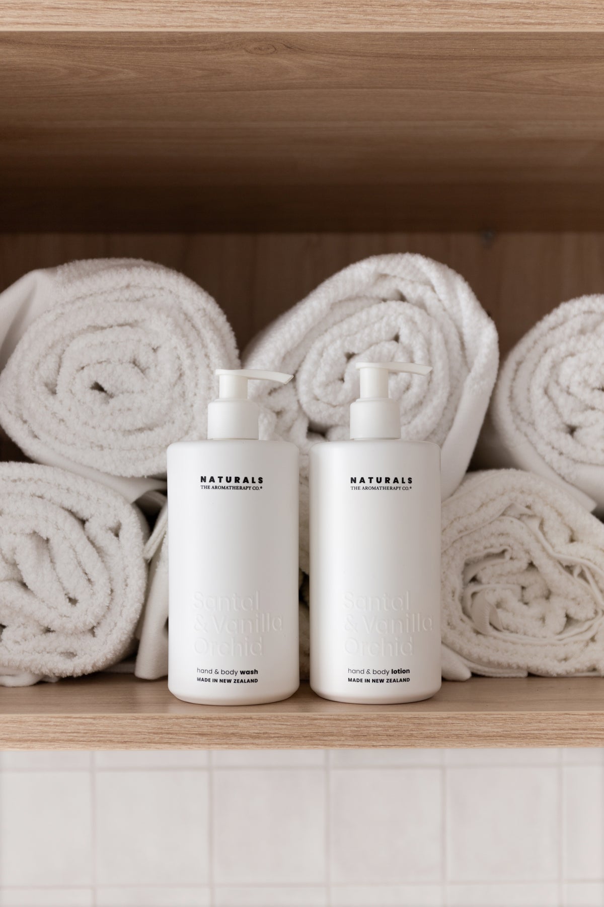 Two Naturals pump bottles on a timber shelf with rolled white towels stacked behind them