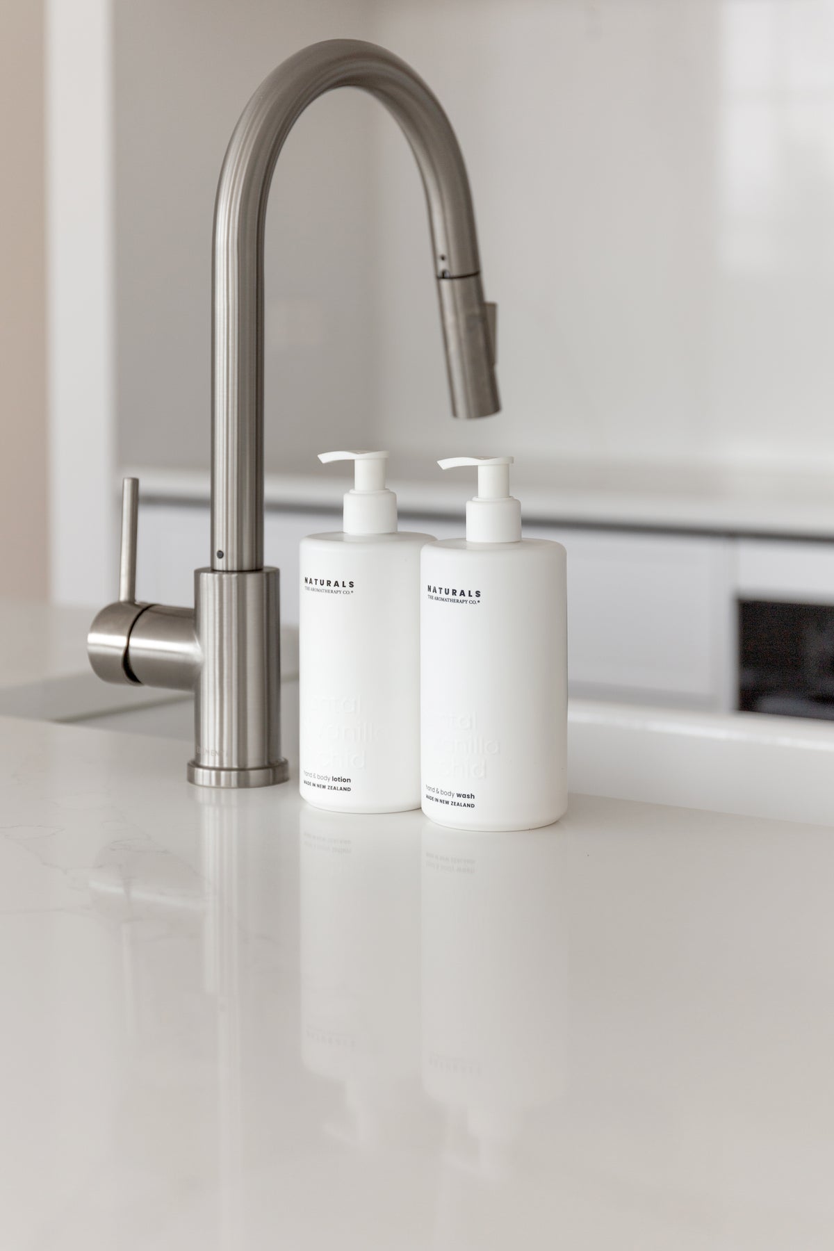 Two Naturals pump bottles on a bright white bench, brushed tapware rising behind them
