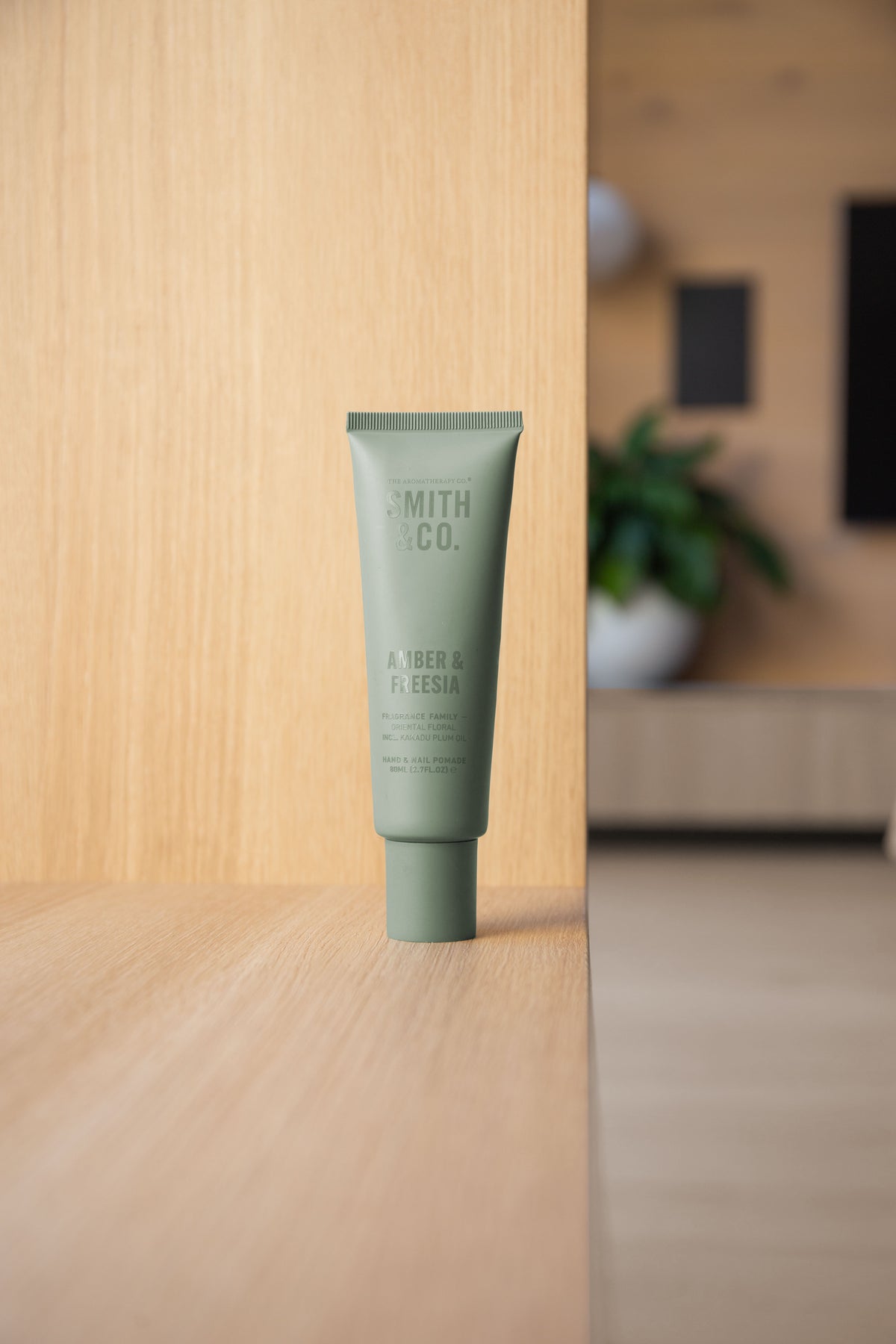 Sage green hand cream tube at the edge of an oak shelf with a living room blurred behind