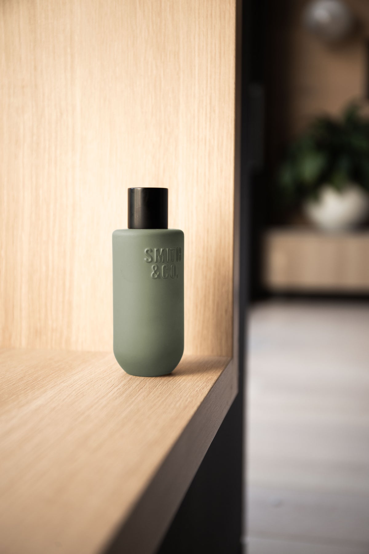 Sage green room spray standing in the corner of an oak shelf beside a dark doorway