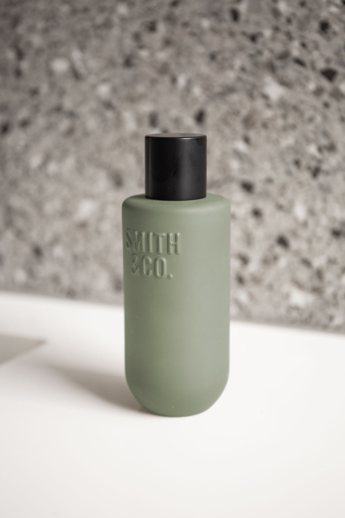 Low angle close up of the sage green bottle and black cap on white against terrazzo