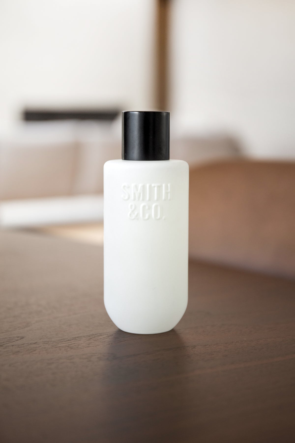 Frosted room spray on a dark timber tabletop with a bedroom blurred in the background