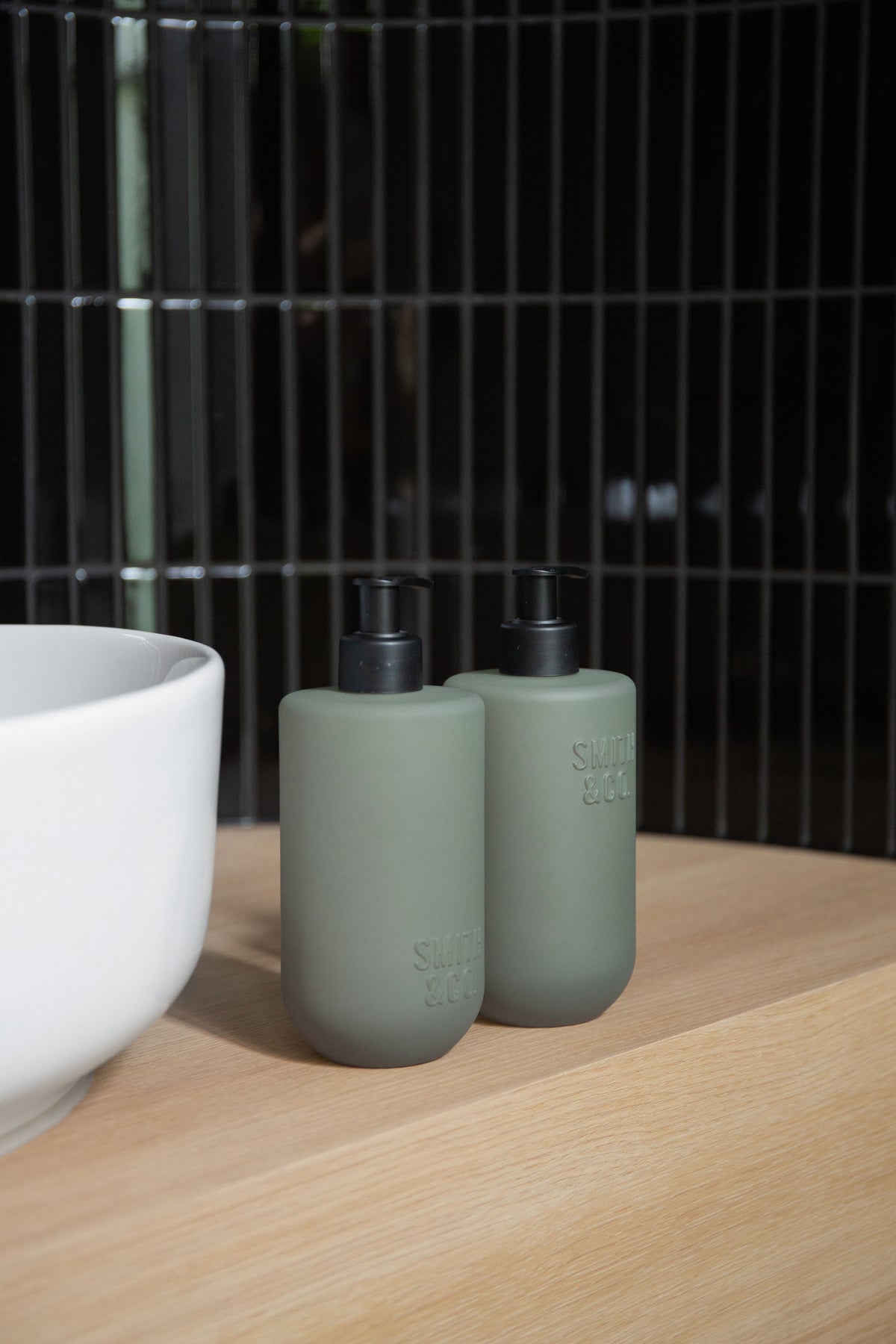 Sage green bottles by a bath against dark green vertical tiles