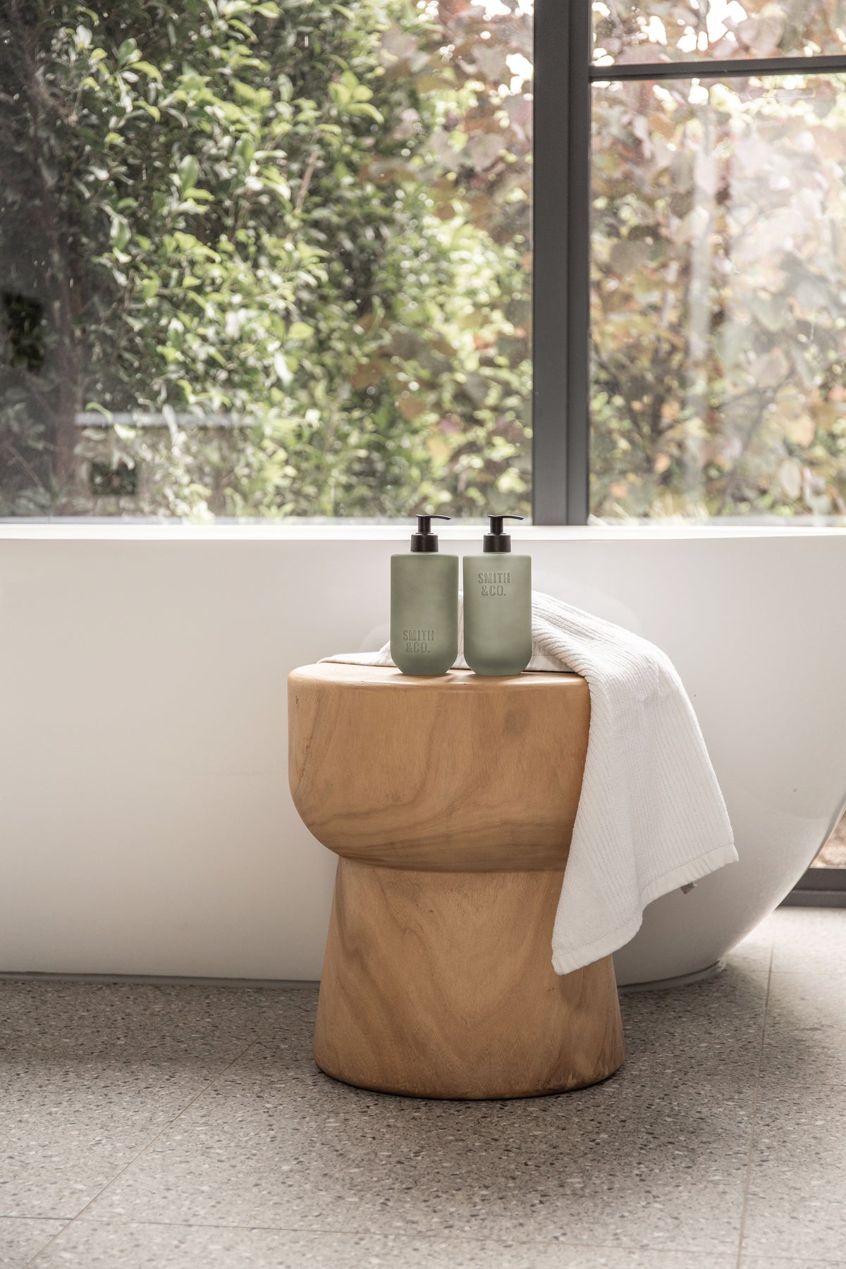 Sage green bottle on a timber stool by a bath and a large window
