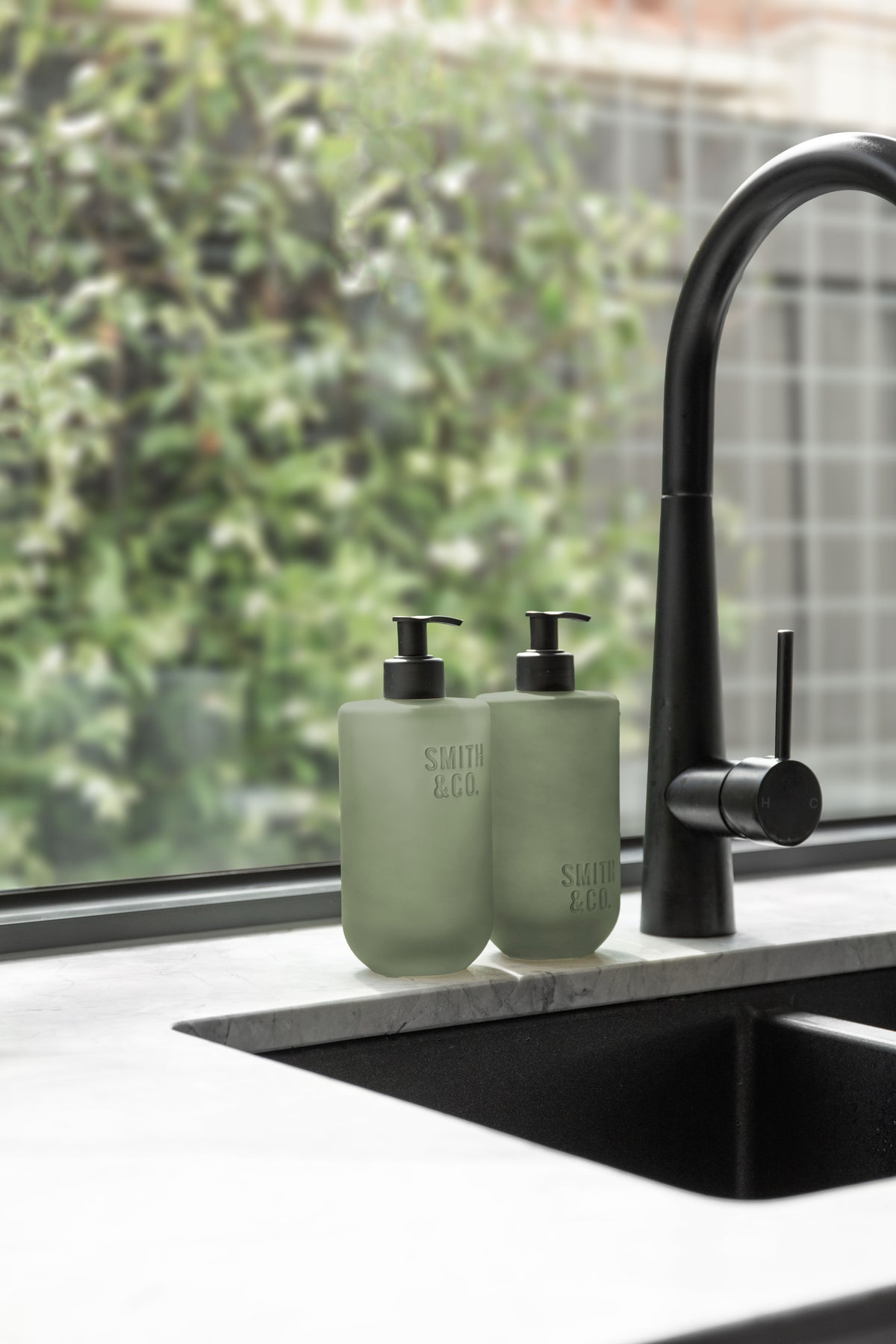 Sage green Smith & Co. bottles at a black kitchen sink with greenery through the window