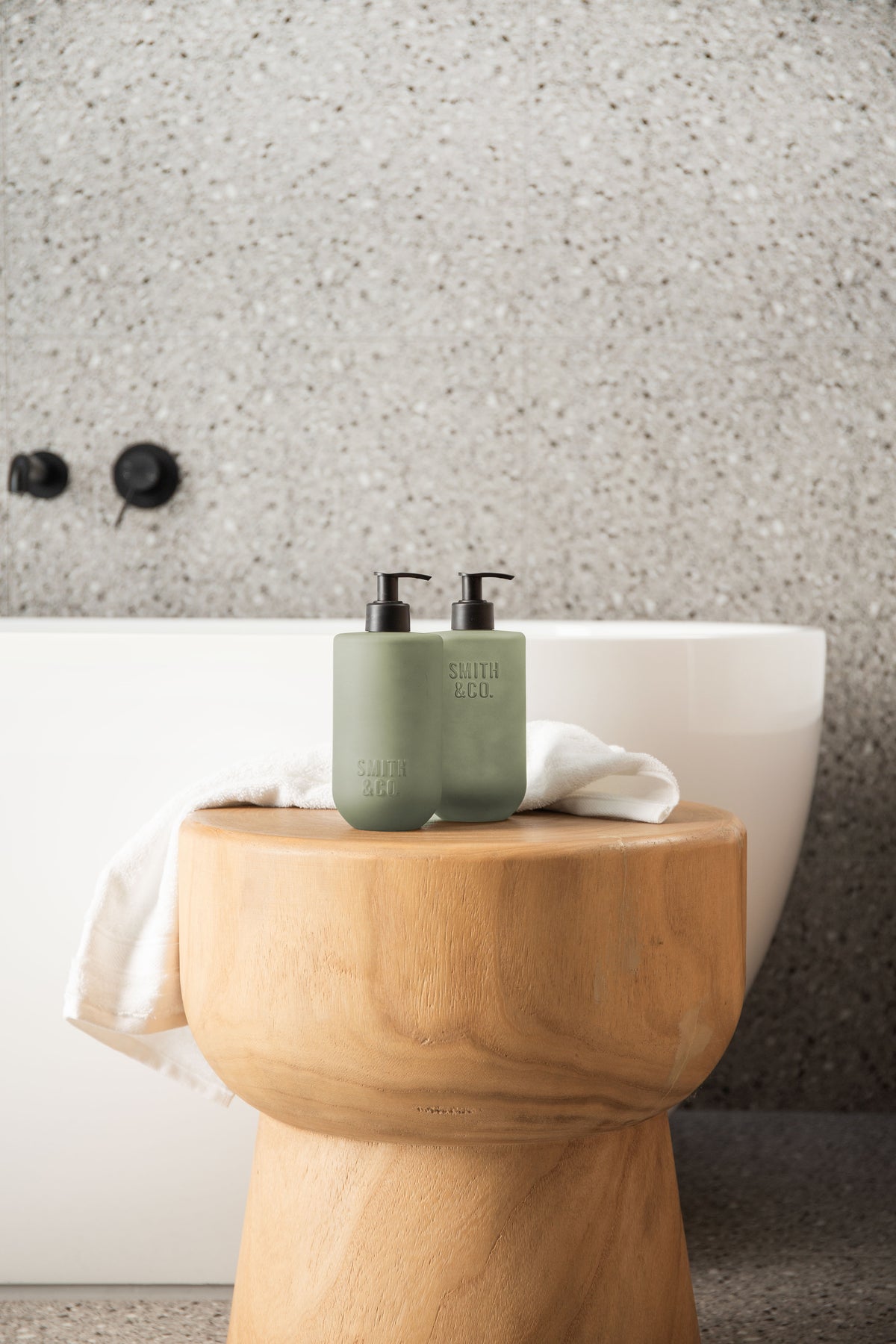 Two sage green bottles on a timber stool against terrazzo walls