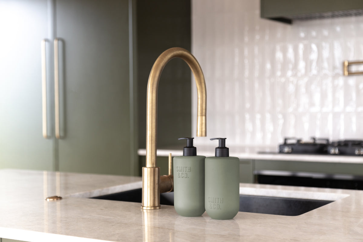 Sage green bottles beside a brass tap and sink on a stone benchtop