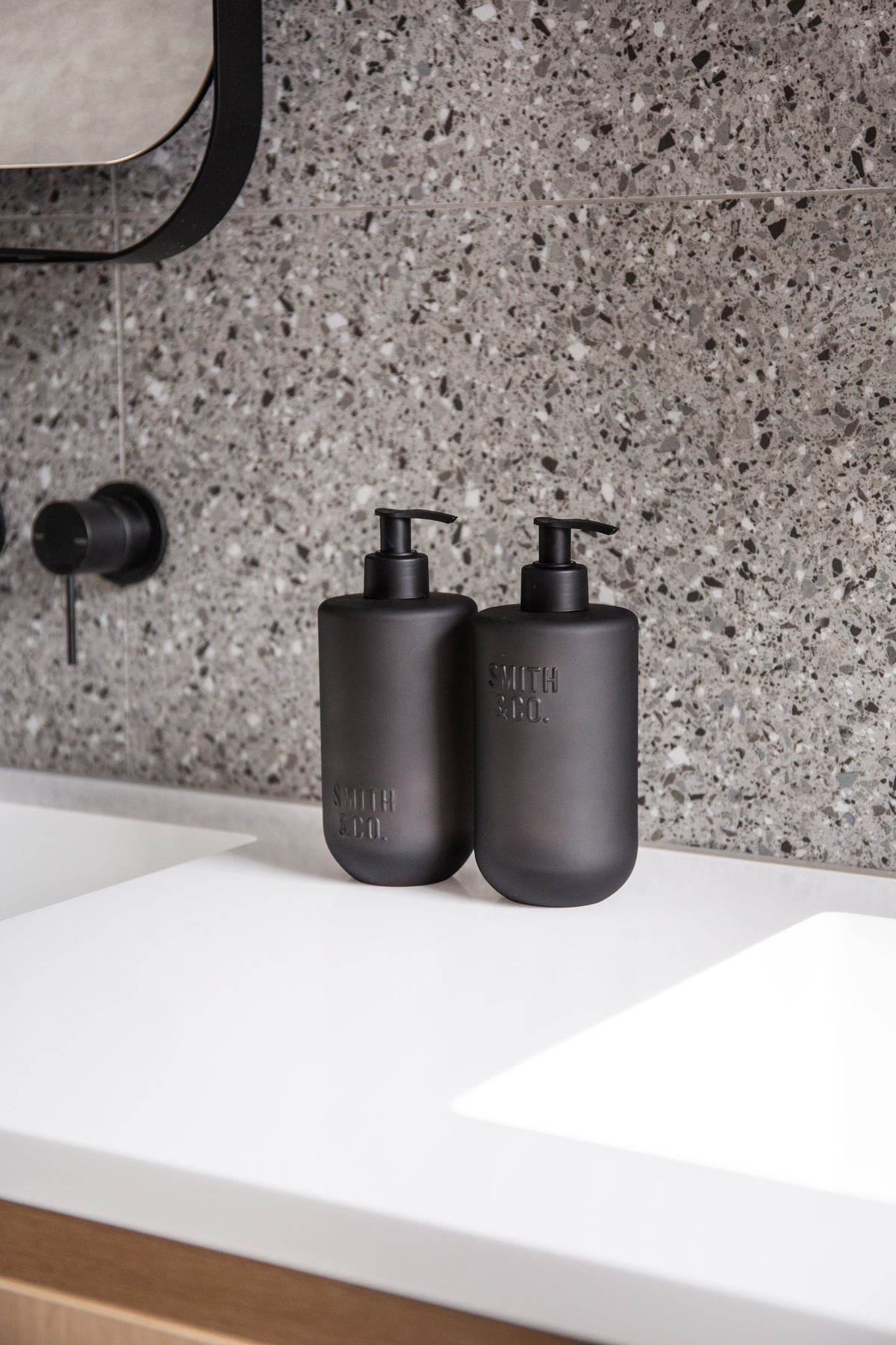 Two Smith & Co. bottles on a white vanity below a round mirror, against terrazzo