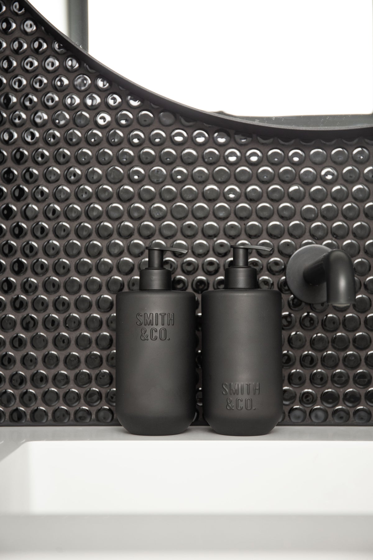 Two Smith & Co. bottles on a white basin against black penny round tiles