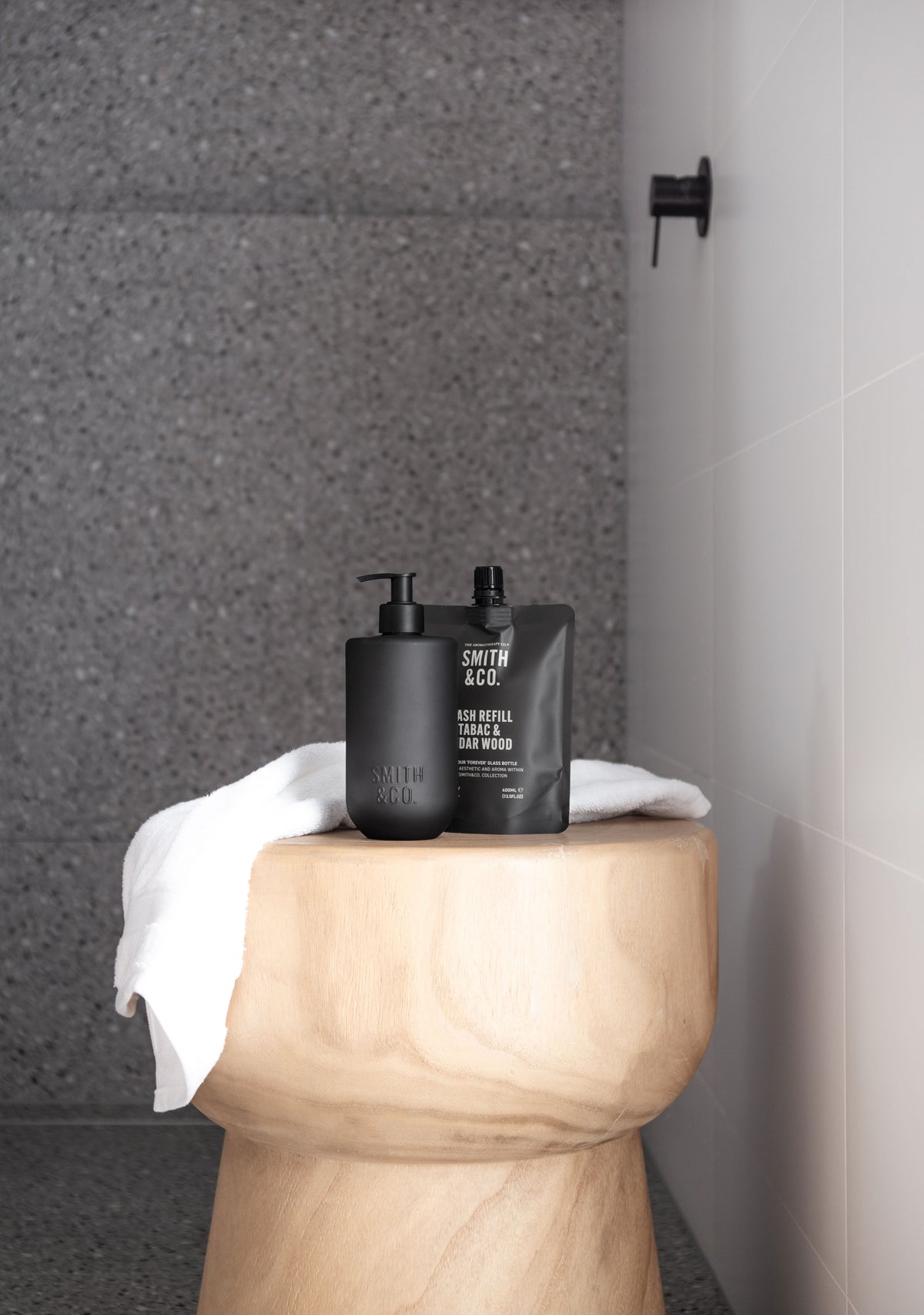 Smith & Co. bottle and pouch on a timber stool with a towel in a grey shower