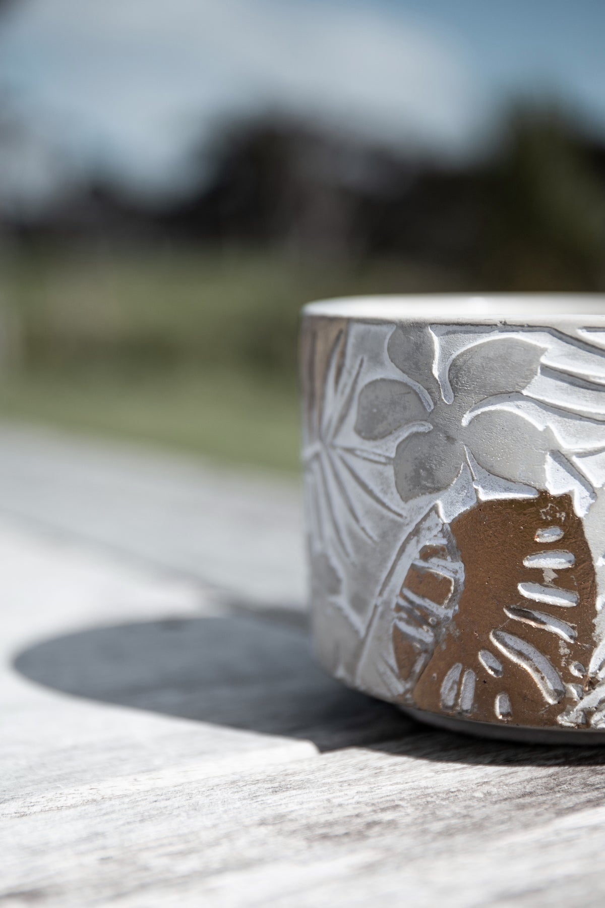 Close crop of the candle's copper and white leaf pattern on a timber bench, countryside blurred behind