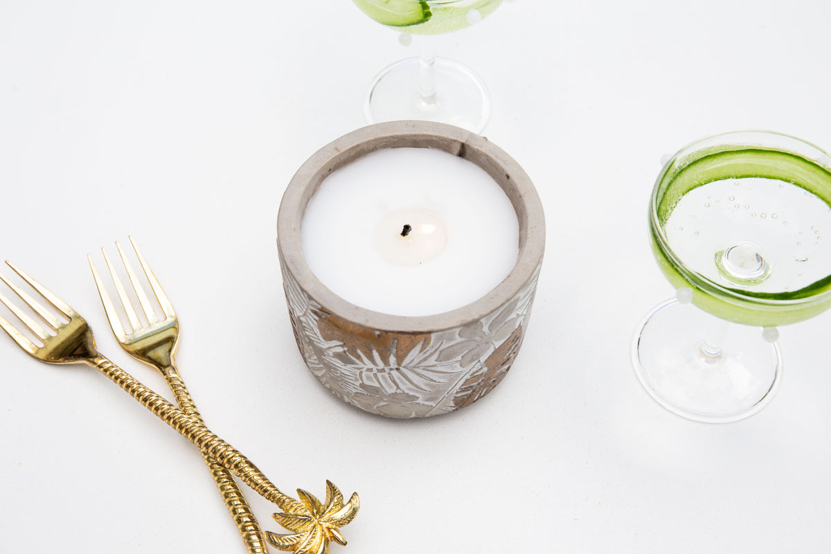 Lit candle from above on white with gold palm-handled forks and a cocktail glass with cucumber