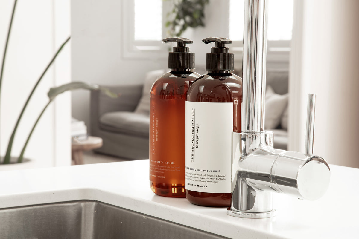 Amber pump bottles beside a kitchen sink and tall chrome tap, a living room soft in the background