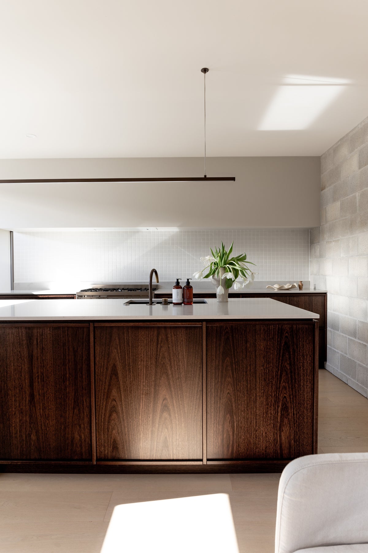 Wide view down a dark timber kitchen island, amber pump bottles and white tulips by the sink