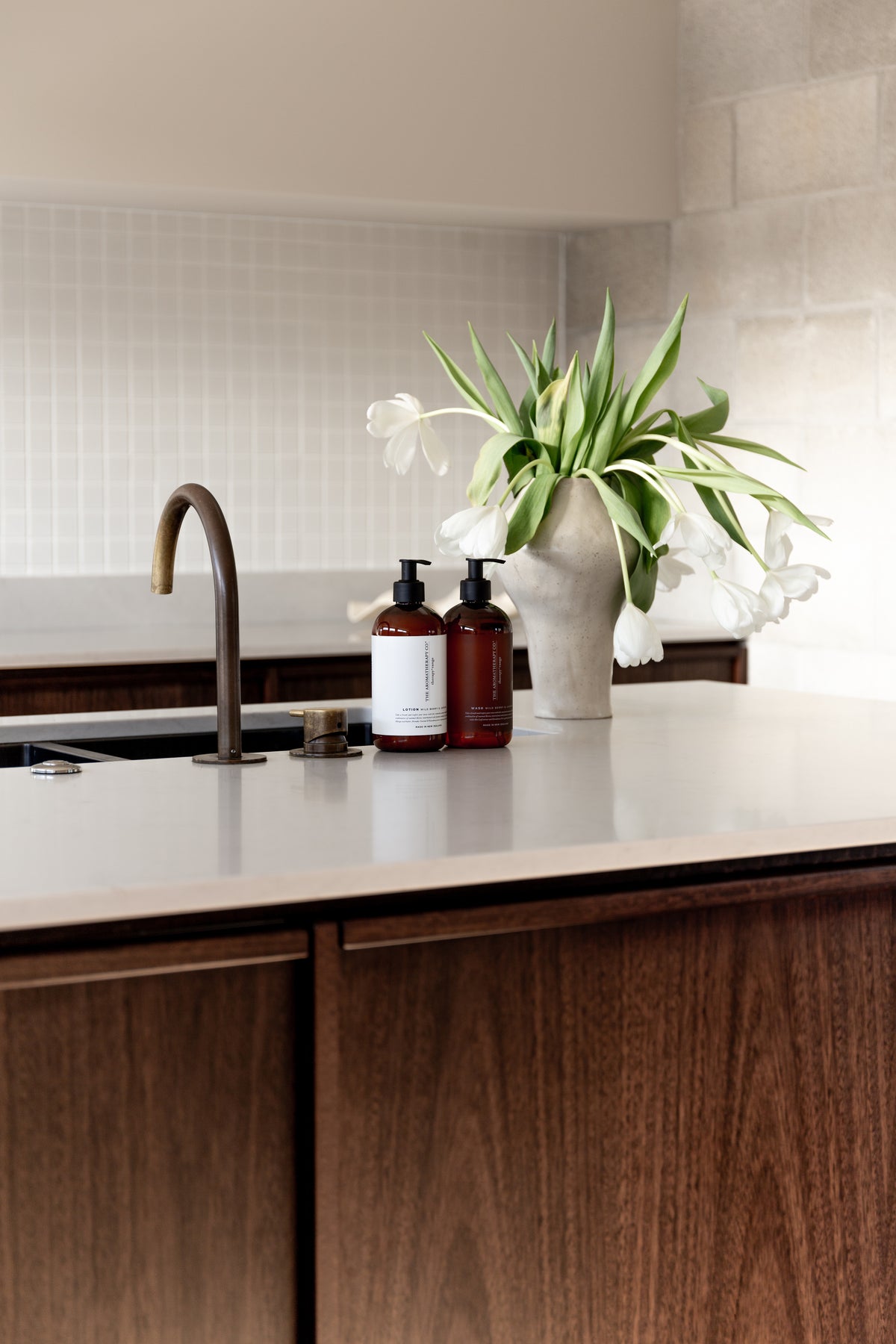 Kitchen island in dark timber with white tulips and amber pump bottles beside the tapware