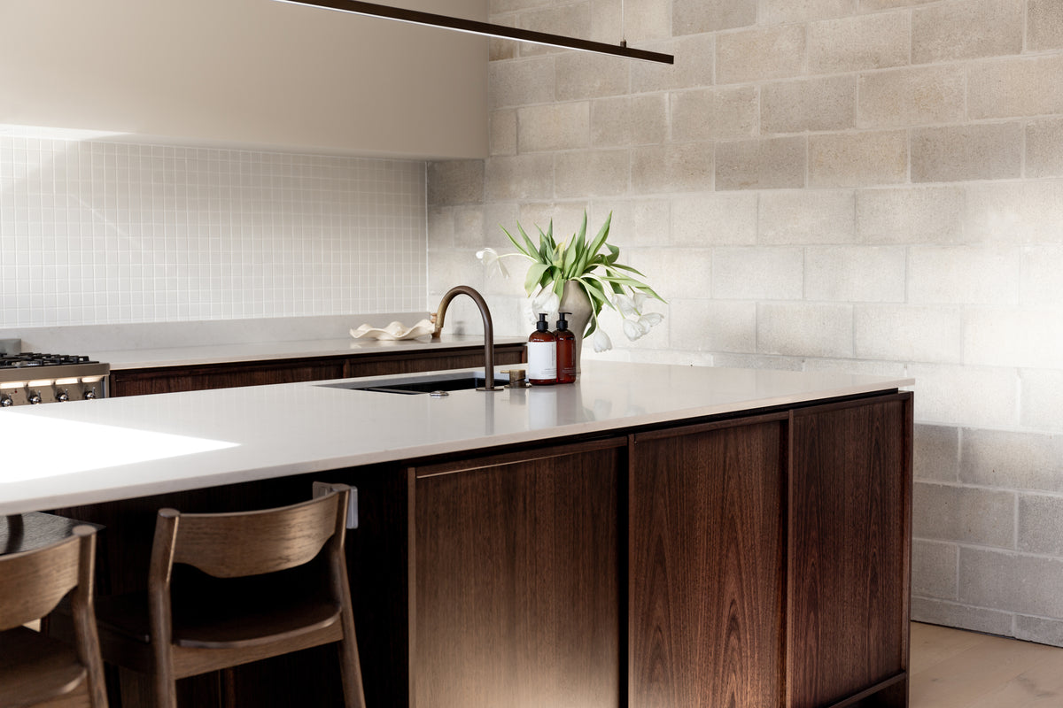 Long dark timber island with a tiled splashback, white tulips and amber bottles at the sink